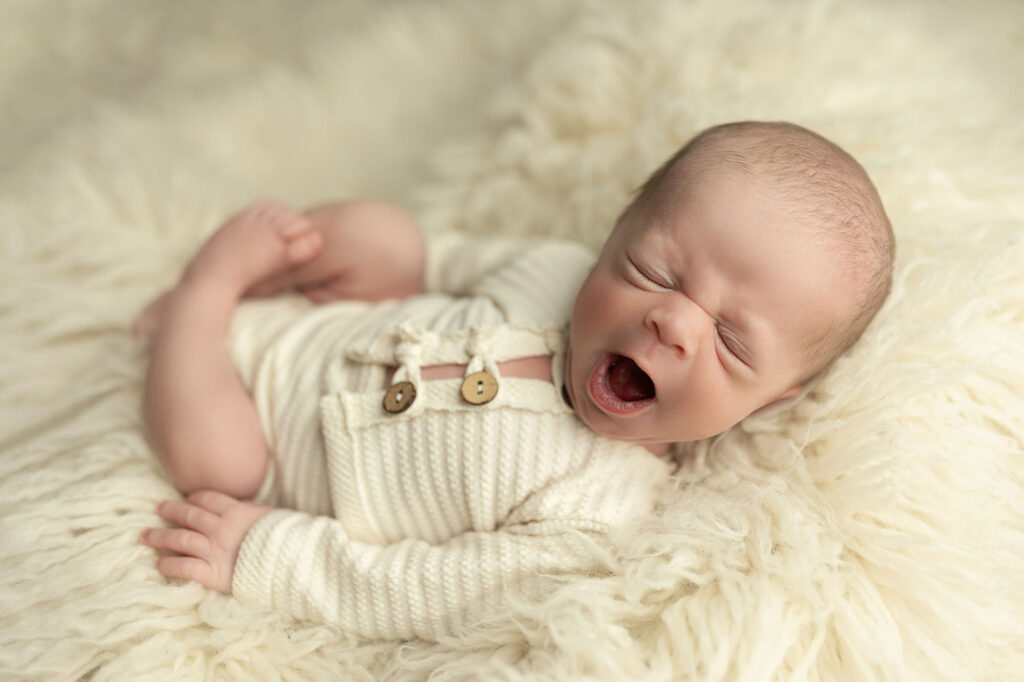 Yawning newborn in a cream knitted outfit, lying on a soft cream rug.